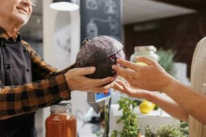 Older man at checkout counter in zero waste shop selling farm grown cabbage to vegan customers with green living lifestyle. Hipster couple buying groceries in local neighborhood store, close up photo