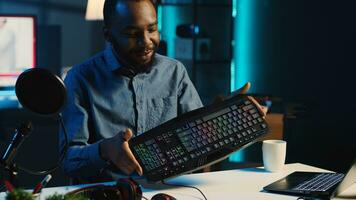Content creator in dimly lit living room uses camera to film keyboard, mouse and headphones review for online streaming platforms. Media star hosts internet show, unboxing wired gaming peripherals photo