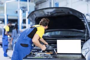 Mockup laptop on car service work station bench with technician in blurry background replacing engine. Isolated screen device next to qualified garage expert fixing client automobile photo