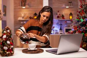 Modern woman filling mug of tea from kettle on video call connection talking to friends. Cheerful young person using laptop for wishing merry christmas in seasonal decorated kitchen photo
