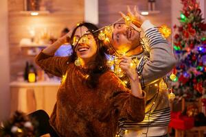 Amused couple caughting in christmas tree light during christmastime preparing xmas decoration for kitchen. Happy funny family enjoying spending winter holiday together photo