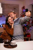 sonriente familia tomando selfie utilizando teléfono inteligente disfrutando Navidad hora en pie en Navidad cocina. contento alegre familia celebrando invierno fiesta estación. nuevo año festivo temporada foto
