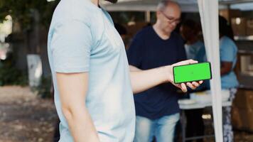 Detailed view of person horizontally holding mobile phone at charitable food drive, presenting an isolated mockup template. Volunteer grasping smartphone with blank green screen display. Tripod shot. photo