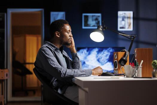 African american man yawning while doing remote work at home and trying to solve tasks before deadline, financial data report. Male employee feeling overworked and dealing with burnout. photo