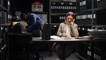 Caucasian private investigator answering phone call, asking about forensic evidence report and reviews documents. Female inspector looks at case files and uses landline to verify private information. photo