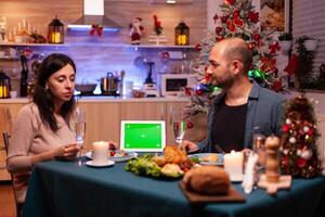 alegre familia disfrutando Navidad cena en Navidad decorado cocina celebrando Navidad día festivo. verde pantalla burlarse de arriba croma llave tableta computadora con aislado monitor en pie en mesa foto