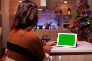 Caucasian adult watching green screen technology on tablet sitting at kitchen counter. Young woman using digital chroma key concept for background template, mockup gadget app on display photo