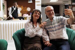 Elderly couple uses a mobile device to make a video chat while on vacation in a fancy hotel lobby. Retired senior travellers relax in the lounge area, talking with relatives on smartphone. photo