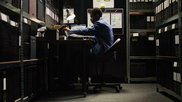 African American detective in archive room, reviewing evidence and files, with reliance on technology. Police officer conducts forensic investigation and background checks, ensuring justice shines. photo