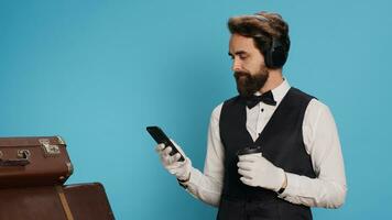 Doorman texting messages drinking coffee in studio, enjoying break with music on headphones. Young adult with classy hotel sector job messaging and sipping on cold brew beverage. photo