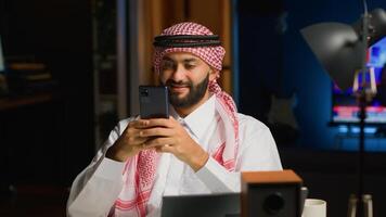 Happy arab man at home in living room typing messages on smartphone. Middle Eastern person holding mobile phone, enjoying relaxing leisure time talking with friends over online messaging app photo