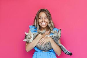 A young caucasian pretty cute blonde woman in a blue dress holds a cat in her hands like a baby isolated on a bright color pink background. The girl babying with a kitten. Fiendship of pet and owner photo