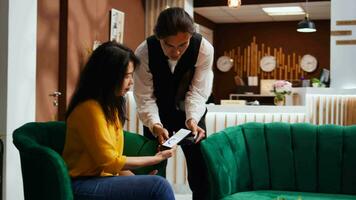 Tourist using nfc payment to buy coffee cup from hotel bar, employee bringing drink in lounge area. Asian woman relaxing in lobby, making mobile phone transaction at pos terminal. Handheld shot. video