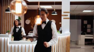 Asian waiter bringing coffee to guest in lounge area, waiting to be called at front desk for room check in. Woman arriving at resort and relaxing with drink before registration, friendly staff. video