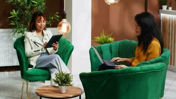 Two friends searching places to visit during their vacation abroad, using laptop and tablet while they wait for registration in lounge area. Asian women passing time with devices. video