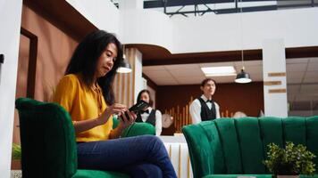 turista desplazamiento social medios de comunicación aplicaciones en teléfono inteligente, utilizando gratis Wifi conexión en salón zona a pasar hora antes de haciendo cheque en. asiático mujer relajante en sofá y planificación viaje itinerario. video