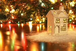 Small ceramic white house candle holder lantern in decorative snow on table, Christmas tree with garland lights on background. Beautiful warm Xmas theme, New Year, winter time concept. photo