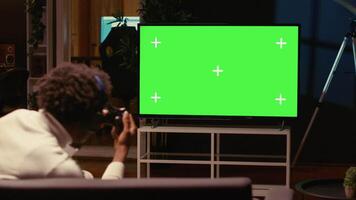 uomo nel debolmente illuminato appartamento giocando video Giochi su verde schermo widescreen inteligente tv, godendo giorno via a partire dal opera. gamer combattendo nemici nel in linea multiplayer tiratore su gioco consolle