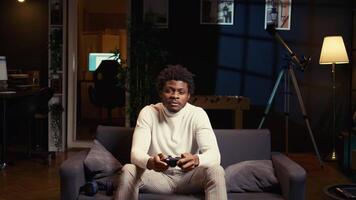 Man focused on playing singleplayer videogames on TV in cozy living room. BIPOC gamer using controller to beat science fiction shooter game on gaming console, relaxing on couch video