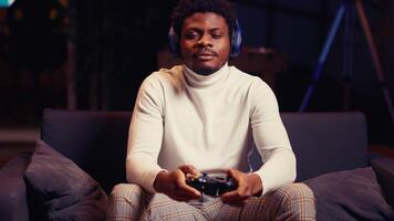 African american man having online gaming session with friends, talking with them through headphones microphone. Gamer playing videogames with teammates on console using gamepad controller video