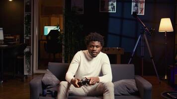 African american man playing shooter videogame in cozy living room, holding controller. Gamer participating in PvP online multiplayer game using console system, relaxing at home video