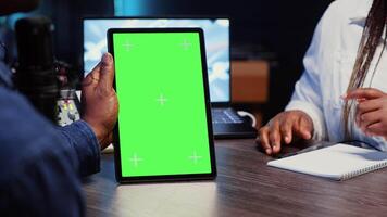 Green screen tablet held by vlogger show host chatting with guest in neon lights ornate living room studio. Close up shot of mockup device used by man and woman broadcasting discussion on podcast video