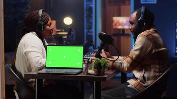 Isolated screen laptop next to vlogger show presenter and guest talking in neon lights ornate studio. Chroma key notebook next to man and woman broadcasting discussion on online podcast video