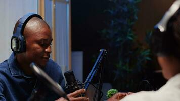 africano americano uomo fare Podcast nel studio, indossare cuffie e parlando su professionale microfono con ospite. vicino su tiro di bipoc soddisfare Creatore intervistando un altro persona video