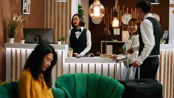 Asian client arriving at reception desk with trolley bags, staff asking tourist about her room reservation for check in process. Hotel concierge greeting guest at holiday resort, tourism concept. video