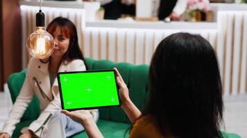 Traveler checking isolated greenscreen template on tablet, sitting with her friend in lounge area and looking at blank copyspace template. Asian guest using isolated mockup. Tripod shot. video