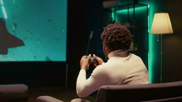 afrikansk amerikan man använder sig av moln gaming service till spela krävande vetenskap fiktion TV-spel på stor TV visa. gamer njuter hög kvalitet grafik, strömning spel uppkopplad, zoom ut skott video