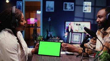 Woman and man participating in podcast, laughing while watching funny internet clips on green screen tablet in studio, discussing content. Podcasters reacting to amusing videos on chroma key device