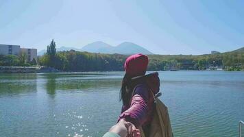 portrait de une fille dans une veste et chapeau sur le jetée avec une magnifique Contexte de Lac et montagnes, suivre moi. une femme points à un inspirant Naturel paysage. video