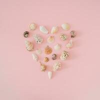 Creative summer layout with love heart symbol made with sea shells on light pastel pink background. Minimal summer concept. Trendy summertime love idea. Sea shells aesthetic. Flat lay. Top of view. photo