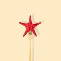 Red starfish pricked on a golden fork on light pastel cream background. Minimal summer concept. Creative trendy sea star with fork idea. Summer aesthetic. photo