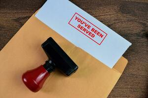 Red Handle Rubber Stamper and You've Been Served text above brown envelope isolated on wooden background photo