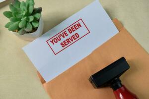 Red Handle Rubber Stamper and You've been served text above Brown envelope isolated on wooden background photo