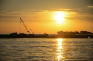 silueta construcción barco y grúas ajuste en marina zona a contrato exportar tubería negocio debajo a mar con hermosa puesta de sol cielo antecedentes. foto