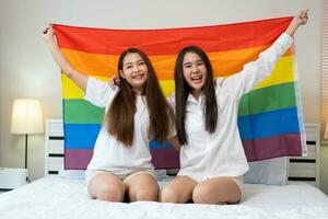 Portraits of happy Asian lesbian couple sitting with rainbow flag sitting bed beside the window. Cheerful two young adult women living together with relationship concept. LGBTQ Lifestyle Pride Month photo