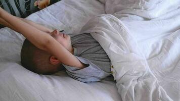 cute eight year old boy wakes up in the morning at home in bed with white linens, rubs his eyes, yawns, smiles, stretches after sleep, healthy sleep for children. video