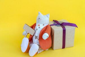 A soft toy cat with a wooden sign with a place for text and a gift box on a yellow background. photo