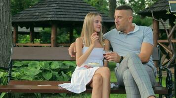 An elderly man and a young girl are sitting together on a park bench. A man drinks a drink in a glass and a girl feeds fruit video