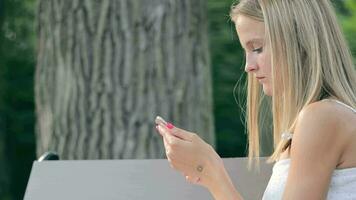 uma jovem caucasiano menina é digitando uma mensagem em uma Smartphone enquanto sentado em uma Banco dentro a verão. lado Visão do uma menina digitando texto em uma telefone dentro a parque. video