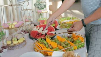 une homme prend fruit de le banquet table et met il sur une plaque. pastèque les raisins et tranché Orange sur buffet tableau. video