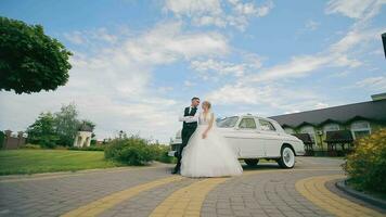 The newlyweds stand near a retro car and kiss against the background of the blue sky. The wedding day of the newlyweds in love, a camera runs over them, they pose for the camera. video