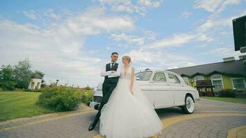 The newlyweds stand near a retro car and kiss against the background of the blue sky. The wedding day of the newlyweds in love, a camera runs over them, they pose for the camera. video