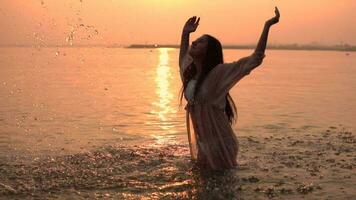 beautiful girl with long hair makes a spray of hands standing in the water against the background of the sun having a good mood. slow motion. HD video