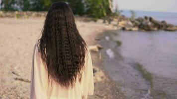 A beautiful girl with long dark hair in a beach tunic walks along the beach with a good mood and turns around. slow motion video