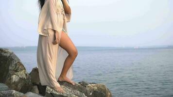 sexig flicka modell med lång hår och i en strand tunika är stående på stenar mot de bakgrund av de hav och Framställ. långsam rörelse. hd video