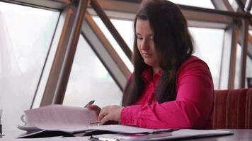bedrijf vrouw met donker lang haar- in helder kleren vult kantoor documenten terwijl zittend Bij tafel in kantoor video